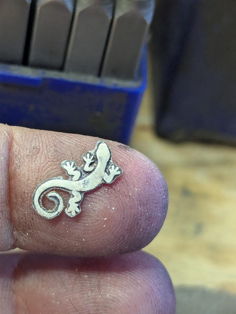 May include: A small, silver-colored lizard pendant with detailed features. The lizard has a curled tail and is resting on a finger. The background includes a blue container and metal tools.
