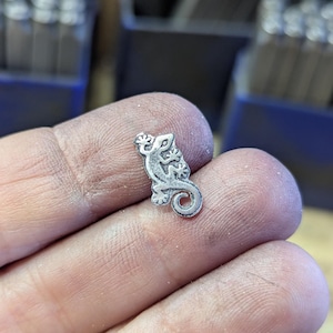 Może przedstawiać: Mały, srebrny metalowy charms w kształcie gekona. Charms jest szczeg&oacute;łowy z wytrawionymi liniami i ma zakręcony ogon. Gekon jest trzymany w dłoni, z innymi narzędziami widocznymi w tle.