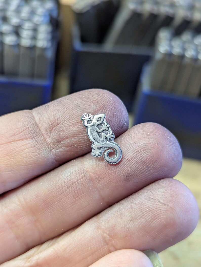 Może przedstawiać: Mały, srebrny metalowy charms w kształcie gekona. Charms jest szczeg&oacute;łowy z wytrawionymi liniami i ma zakręcony ogon. Gekon jest trzymany w dłoni, z innymi narzędziami widocznymi w tle.