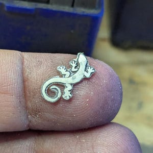 May include: A small, silver-colored lizard charm with detailed features. The charm is held between fingers, showcasing its intricate design. The lizard has a curled tail and detailed feet, suggesting a jewelry or craft item.