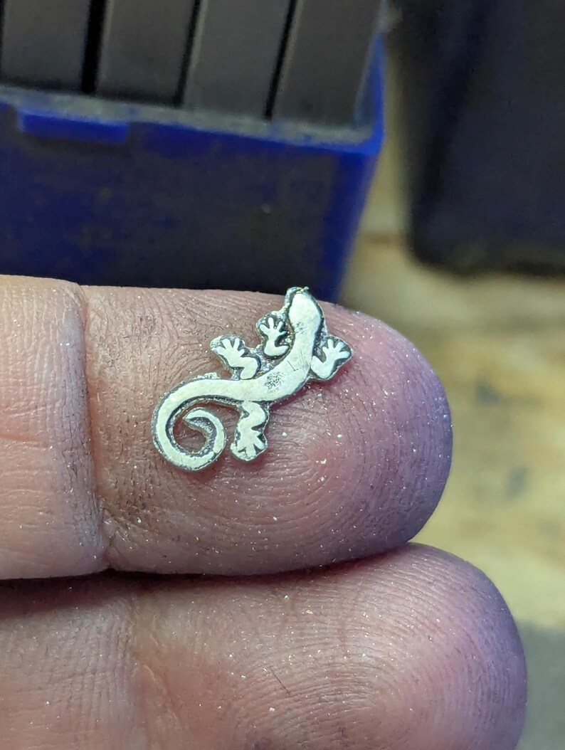 May include: A small, silver-colored lizard charm with detailed features. The charm is held between fingers, showcasing its intricate design. The lizard has a curled tail and detailed feet, suggesting a jewelry or craft item.