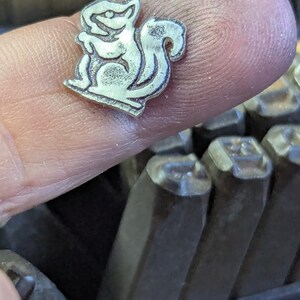 Op de afbeelding: Een kleine, zilverkleurige metalen stempel in de vorm van een eekhoorn. De stempel wordt tussen vingers gehouden, met een set metalen stempels op de achtergrond. De eekhoorn is gestileerd en cartoonachtig afgebeeld, met een volle staart en een rechte houding.