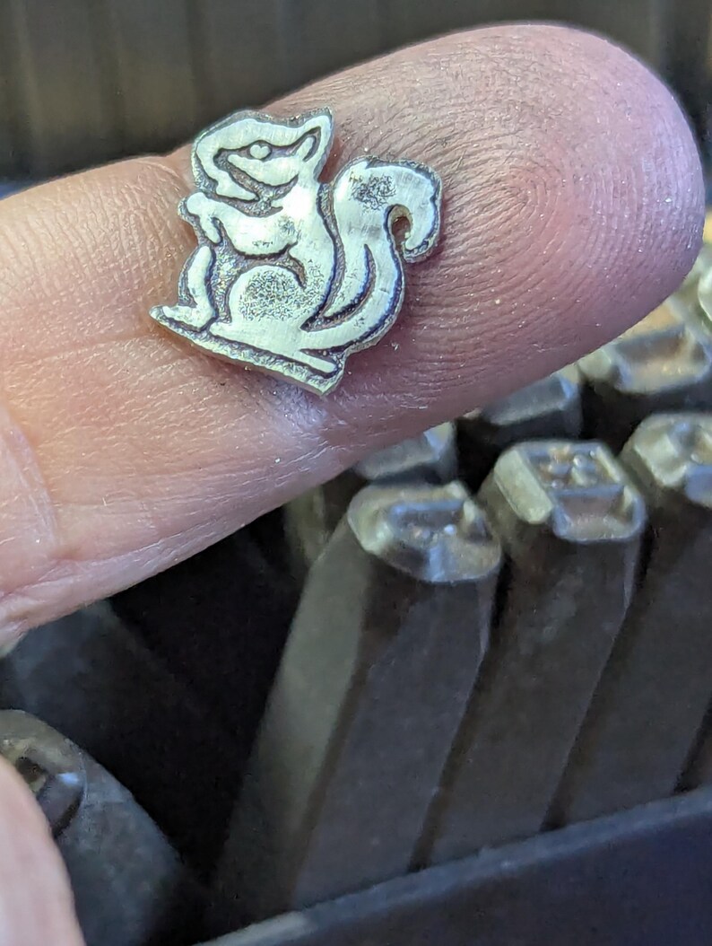 Op de afbeelding: Een kleine, zilverkleurige metalen stempel in de vorm van een eekhoorn. De stempel wordt tussen vingers gehouden, met een set metalen stempels op de achtergrond. De eekhoorn is gestileerd en cartoonachtig afgebeeld, met een volle staart en een rechte houding.