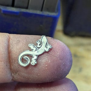 May include: A small, silver-colored lizard charm with detailed features, including tiny feet and a curled tail. The charm is held between fingers, with a blue tool in the background.