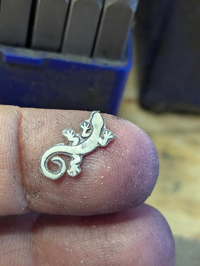 May include: A small, silver-colored lizard charm with detailed features, including tiny feet and a curled tail. The charm is held between fingers, with a blue tool in the background.