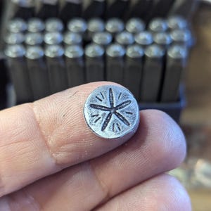 K&ouml;nnte beinhalten: Ein kleiner, kreisf&ouml;rmiger Metallstempel mit einem Sternmuster. Der Stempel wird in den Fingern einer Person gehalten, mit einer Reihe &auml;hnlicher Stempel im Hintergrund. Das Sternmuster hat sechs Punkte und ist in das Metall ge&auml;tzt.