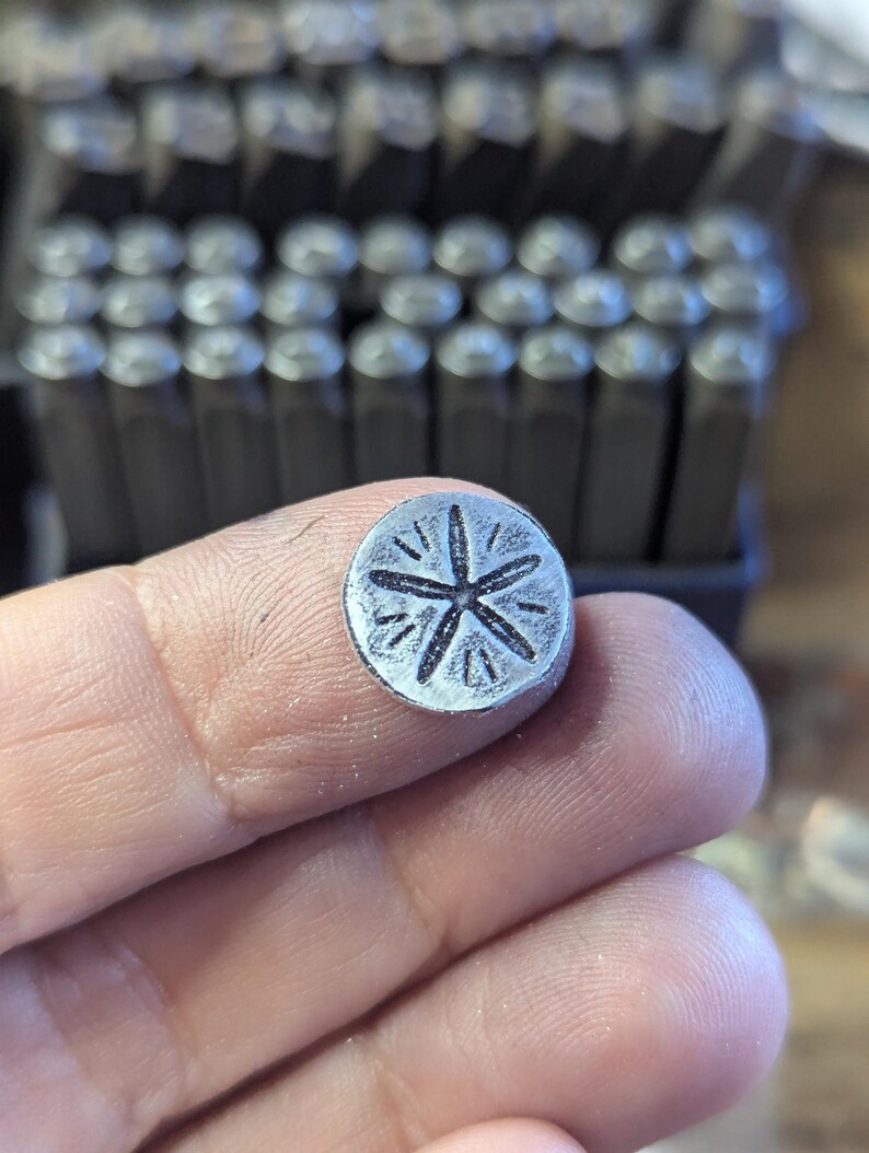 K&ouml;nnte beinhalten: Ein kleiner, kreisf&ouml;rmiger Metallstempel mit einem Sternmuster. Der Stempel wird in den Fingern einer Person gehalten, mit einer Reihe &auml;hnlicher Stempel im Hintergrund. Das Sternmuster hat sechs Punkte und ist in das Metall ge&auml;tzt.