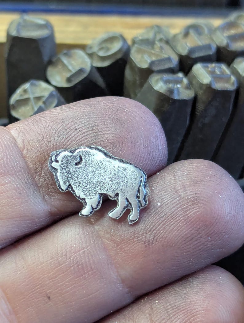 May include: A small, silver-colored metal buffalo charm. The charm is detailed with textured fur and a defined outline. The background shows several metal stamps, suggesting a crafting or jewelry-making context.