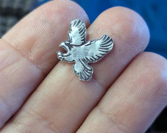 May include: A small, silver-colored eagle charm with outstretched wings and an open beak. The charm is held in a person's hand, showcasing its detailed design. The eagle is a symbol of freedom and power.