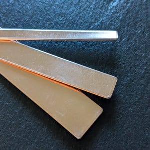 May include: Close-up of several rectangular metal bars with a copper core, arranged on a dark textured surface. The bars have a brushed metal finish, reflecting light. The image highlights the contrast between the metal and the dark background.