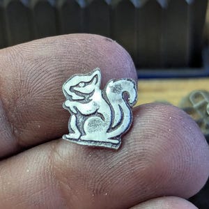 Op de afbeelding: Een kleine, zilverkleurige metalen bedel in de vorm van een eekhoorn. De eekhoorn wordt afgebeeld in een staande houding met zijn staart omhoog gekruld. De bedel wordt tussen vingers gehouden, met een vage achtergrond van metalen gereedschap.
