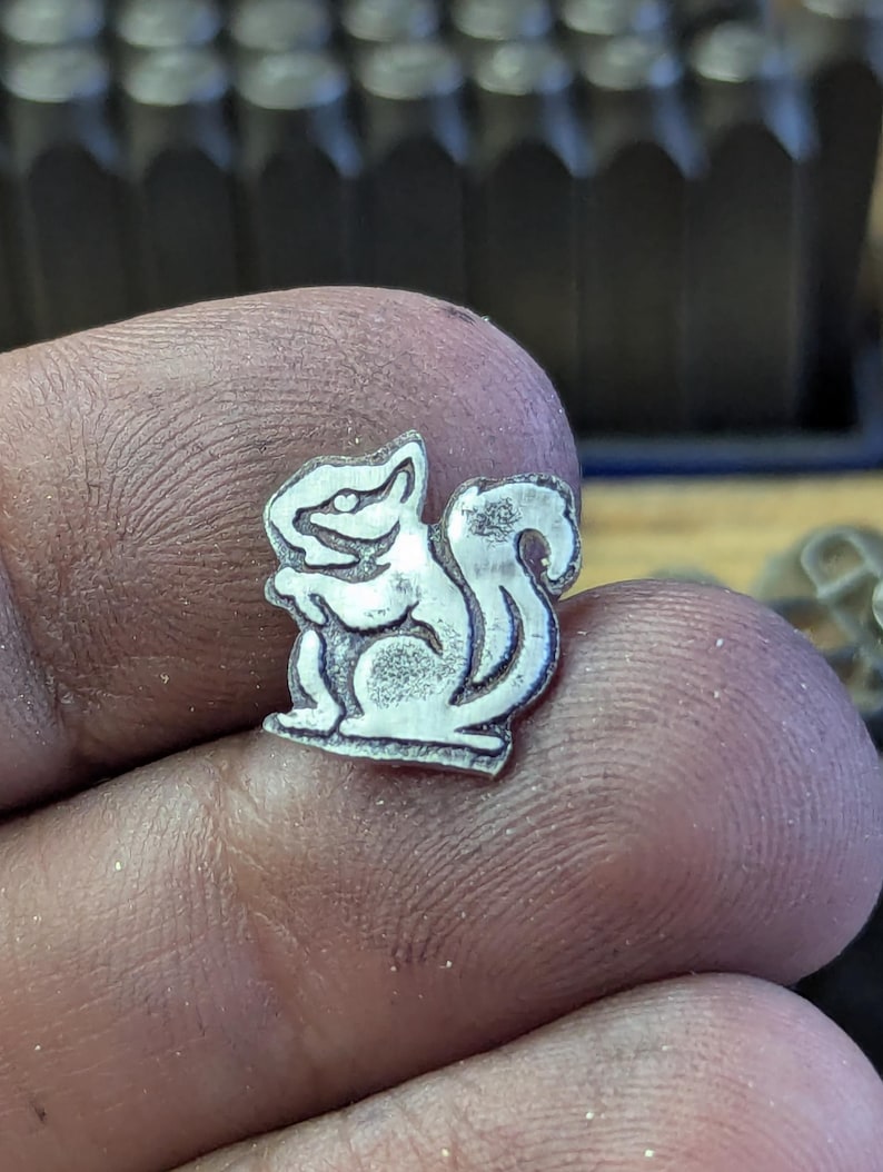 Op de afbeelding: Een kleine, zilverkleurige metalen bedel in de vorm van een eekhoorn. De eekhoorn wordt afgebeeld in een staande houding met zijn staart omhoog gekruld. De bedel wordt tussen vingers gehouden, met een vage achtergrond van metalen gereedschap.