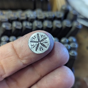 K&ouml;nnte beinhalten: Ein kleiner, kreisf&ouml;rmiger Metallstempel mit einem Sternmuster. Der Stempel wird zwischen den Fingern gehalten, mit einem verschwommenen Hintergrund aus anderen Metallstempeln. Der Stern hat strahlenf&ouml;rmige Linien und wird wahrscheinlich zum Aufpr&auml;gen von Designs verwendet.