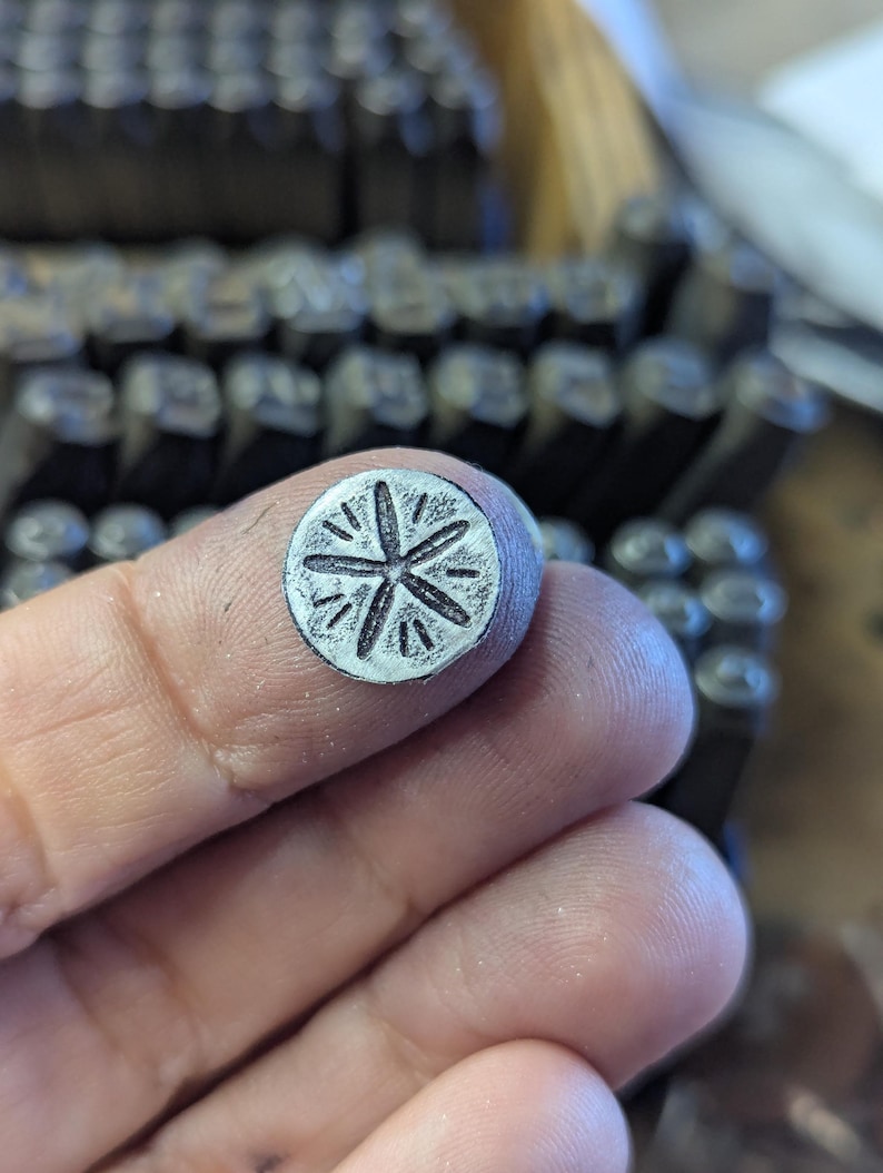 K&ouml;nnte beinhalten: Ein kleiner, kreisf&ouml;rmiger Metallstempel mit einem Sternmuster. Der Stempel wird zwischen den Fingern gehalten, mit einem verschwommenen Hintergrund aus anderen Metallstempeln. Der Stern hat strahlenf&ouml;rmige Linien und wird wahrscheinlich zum Aufpr&auml;gen von Designs verwendet.