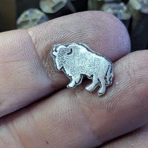 May include: A small, silver-colored metal charm in the shape of a bison. The charm is held in a hand, with other metal tools visible in the background. The bison has detailed features and a textured surface.