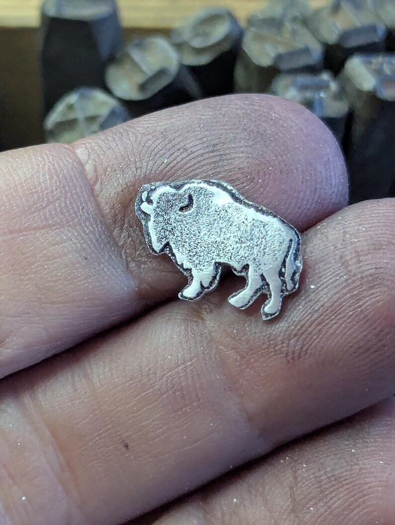 May include: A small, silver-colored metal charm in the shape of a bison. The charm is held in a hand, with other metal tools visible in the background. The bison has detailed features and a textured surface.