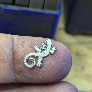 May include: A small, silver-colored lizard charm with detailed features, including tiny feet and a curled tail. The charm is held between fingers, with a blurred background of tools.