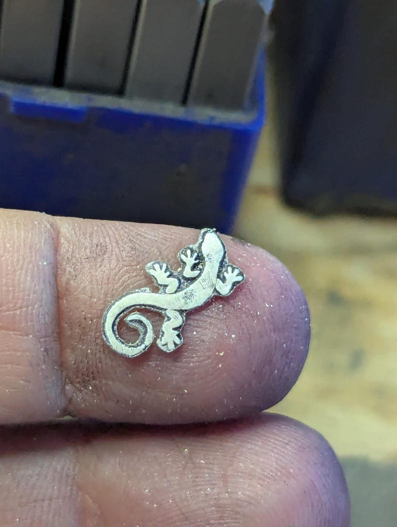 May include: A small, silver-colored lizard charm with detailed features, including tiny feet and a curled tail. The charm is held between fingers, with a blurred background of tools.