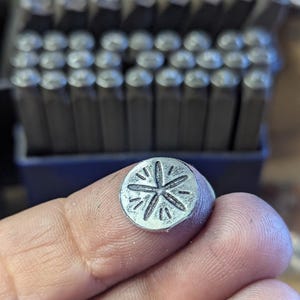 K&ouml;nnte beinhalten: Ein kleiner, runder Metallstempel mit einem Sternmuster, der zwischen Fingern gehalten wird. Der Stempel ist silberfarben und hat ein detailliertes, eingraviertes Muster. Im Hintergrund ist ein Satz Metallstempel zu sehen, was auf ein Werkzeug f&uuml;r die Metallbearbeitung oder das Handwerk hindeutet.