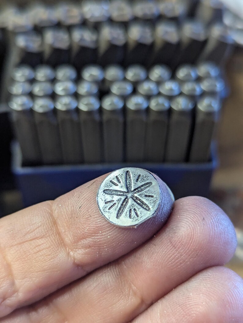 K&ouml;nnte beinhalten: Ein kleiner, runder Metallstempel mit einem Sternmuster, der zwischen Fingern gehalten wird. Der Stempel ist silberfarben und hat ein detailliertes, eingraviertes Muster. Im Hintergrund ist ein Satz Metallstempel zu sehen, was auf ein Werkzeug f&uuml;r die Metallbearbeitung oder das Handwerk hindeutet.