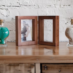 May include: Two heart-shaped glass bottles with cork stoppers, one filled with green sand and the other with white sand, are placed on a wooden surface next to a wooden picture frame with a photo of a couple inside.