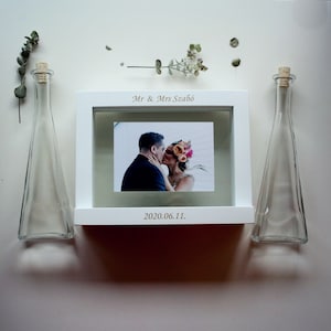 May include: White wooden frame with a photo of a couple kissing. The frame has the text "Mr & Mrs Szabo" and "2020.06.11." on it. Two clear glass bottles with cork stoppers are on either side of the frame.