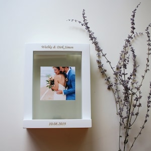 May include: White picture frame with a glass front and a white mount. The frame has a photo of a couple on their wedding day. The frame is engraved with the names "Wiebke & Dirk Simon" and the date "10.08.2019".