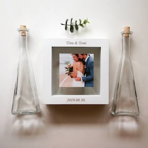 May include: White picture frame with a photo of a couple on their wedding day. The frame has the text "Tina & Tomi" and "2019.08.30."  Two clear glass bottles with cork stoppers are on either side of the frame.