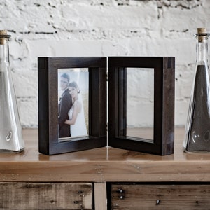 May include: A dark wood picture frame with a glass front, holding a photo of a couple. The frame is open and the photo is visible. Two clear glass bottles with cork tops are on either side of the frame.