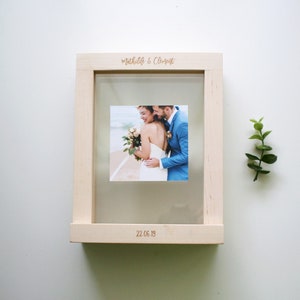 May include: A wooden photo frame with a clear glass insert. The frame has a light wood finish and is engraved with the names "Mathilde & Clement" and the date "22.06.19". A photo of a couple is visible through the glass.
