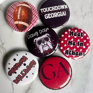 May include: A set of six circular pin-back buttons featuring various designs including a brown football, a bulldog wearing sunglasses, and text such as TOUCHDOWN GEORGIA!, Dawg Days, Meet Me in Athens, GEORGIA BULLDOGS, and GA, with some buttons accented by small beads.