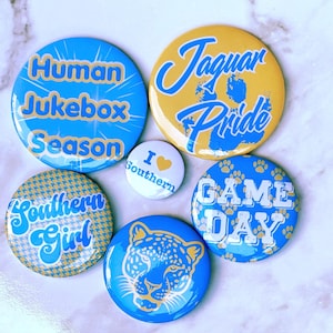 May include: A collection of six circular pin-back buttons in blue and gold. Designs include text reading Human Jukebox Season, Jaguar Pride, I heart Southern, Southern Girl, and Game Day, alongside a graphic of a jaguar face and paw prints.