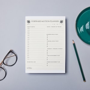 Può includere: Un planner bianco con la scritta "Forward Motion Planner" stampata sulla copertina. Il planner è aperto a una pagina con sezioni per "Today's Motto", "Quote/Thought of the Day", "Business Goals Today", "Personal Goals Today", "Workout/Personal Interest", "Lunch Nourishment Plan" e "Dinner Nourishment Plan". C'è una matita, un paio di occhiali e una tazza di caffè con un piattino verde su una superficie grigia.
