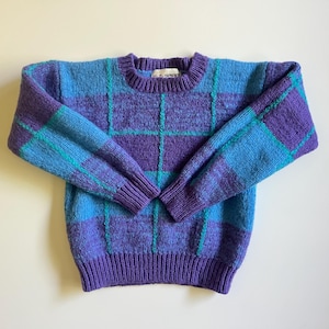 Puede incluir: Un suéter de punto de manga larga con un patrón de cuadros en tonos morado y azul brillante. El diseño incluye líneas finas verticales y horizontales de color turquesa que forman una cuadrícula en la prenda. Tiene cuello redondo acanalado.