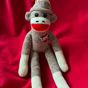 May include: A handmade sock monkey doll with a red mouth, black button eyes, and a red pom-pom on its hat. The monkey is primarily brown and white, with the word "PUNK" on its chest. The background is a solid red.