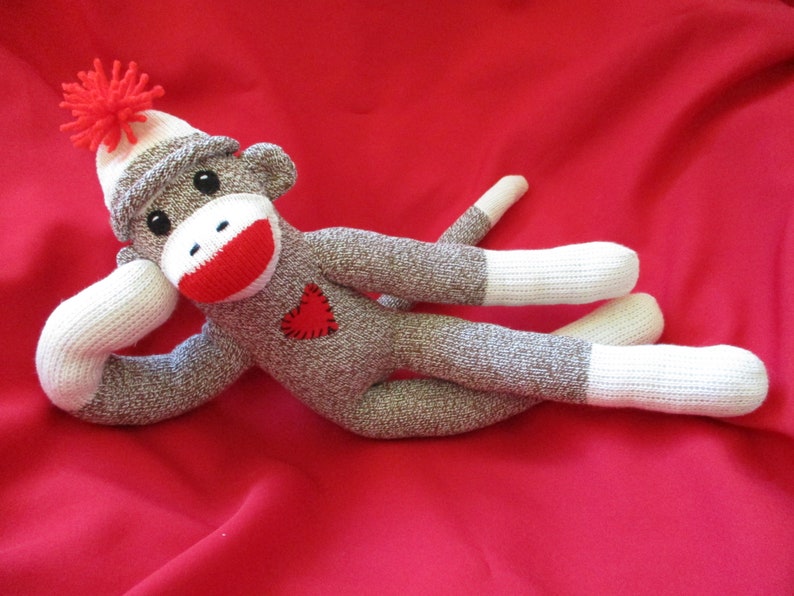 May include: A sock monkey toy with a red heart on its chest, wearing a white knit hat with a red pom-pom on top. The monkey is lying on its side with its arm resting on its head.