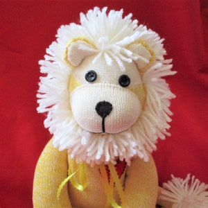 May include: A white and yellow sock monkey lion with a white fluffy mane and black button eyes. The lion is wearing a red heart and a yellow ribbon around its neck.