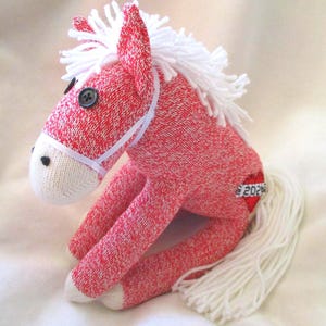 May include: A handmade sock horse in red and white knit fabric. The horse has a white mane and tail, button eyes, and a white bridle. A small tag on the horse's rear reads "2023".
