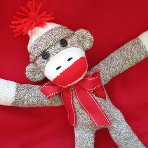May include: A sock monkey with a red hat and a red bow tie with a white heart on it. The monkey is lying on a red background.