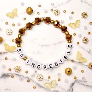 May include: A gold and white beaded bracelet with the words "SO INCREDIBLE" spelled out. The bracelet is surrounded by decorative beads, butterflies, and flowers in gold and white.