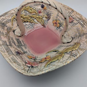 May include: A square, vintage ceramic basket with a pink interior and handle. The basket features a cream-colored exterior with a black grid pattern and gold accents. It is decorated with gold leaves and colorful flowers.