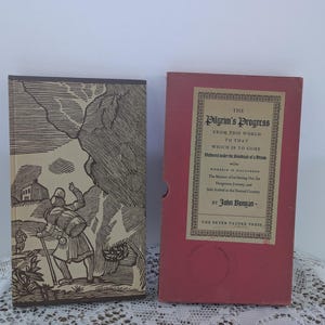 May include: Two vintage books. The first book has a brown cover with a black and white illustration of a person walking with a staff. The second book is in a red case with the title "The Pilgrim's Progress" in gold lettering.