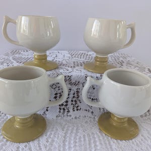 May include: Four cream-colored ceramic mugs with a unique goblet-style design. Each mug features a rounded bowl, a curved handle, and a stem and base in a muted yellow-green hue. The mugs are arranged on a white lace tablecloth.