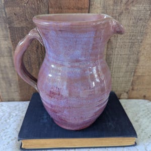 May include: A handmade ceramic pitcher with a rounded body and a curved handle. The pitcher is glazed in shades of purple and mauve, with a glossy finish. It has a spout for easy pouring and is sitting on a dark blue book.