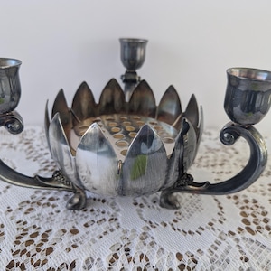 May include: A silver-toned centerpiece with a floral design. It features a central bowl with a grid of holes, flanked by two candle holders and a single central candle holder. The piece has a decorative, ornate design.