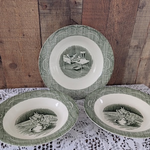 Może przedstawiać: Zestaw trzech talerzy ceramicznych w kolorze zielono-białym w stylu vintage. Talerze mają zieloną obwódkę z dekoracyjnym wzorem i centralną ilustracją. Największy talerz przedstawia scenę z pokoju, a mniejsze miski dekoracyjną urnę.