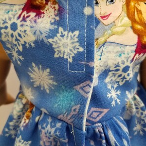 May include: A blue doll dress featuring a snowflake pattern and a portrait of a cartoon character. The dress has a white trim and is made of fabric. The dress is designed for a doll.
