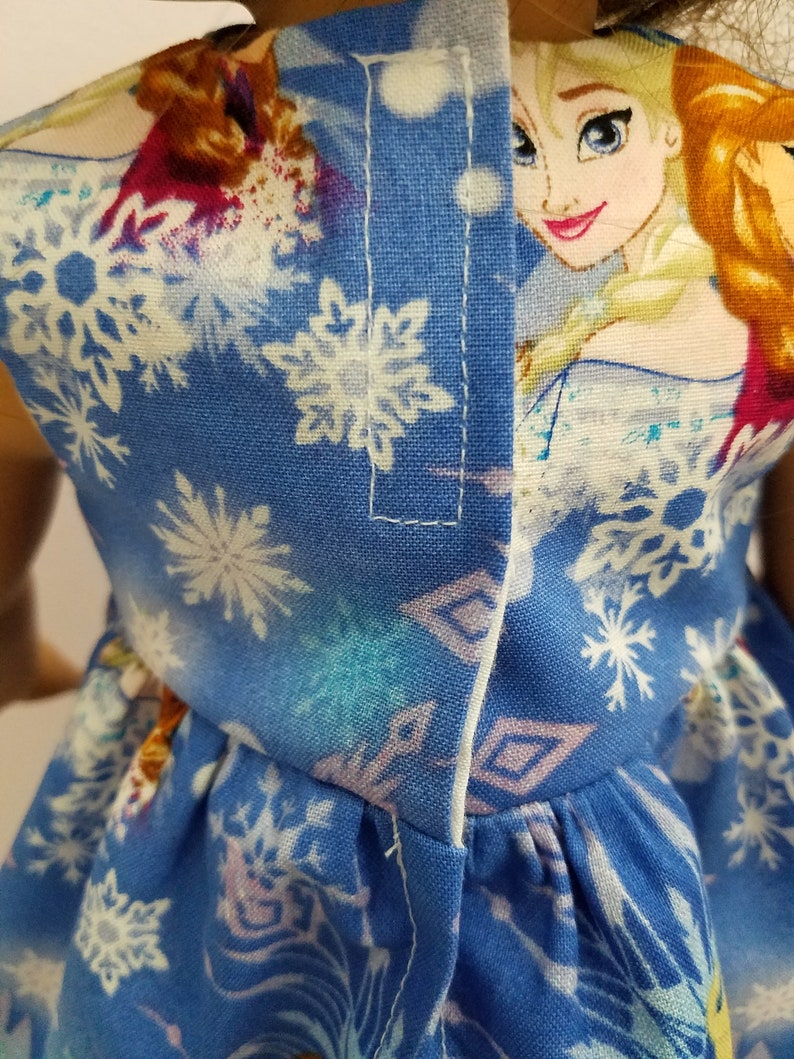May include: A blue doll dress featuring a snowflake pattern and a portrait of a cartoon character. The dress has a white trim and is made of fabric. The dress is designed for a doll.