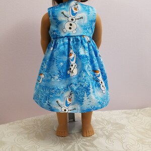 May include: A blue doll's dress with a pattern of Olaf the snowman from Frozen. The dress has a gathered skirt and a light blue and white winter forest background.
