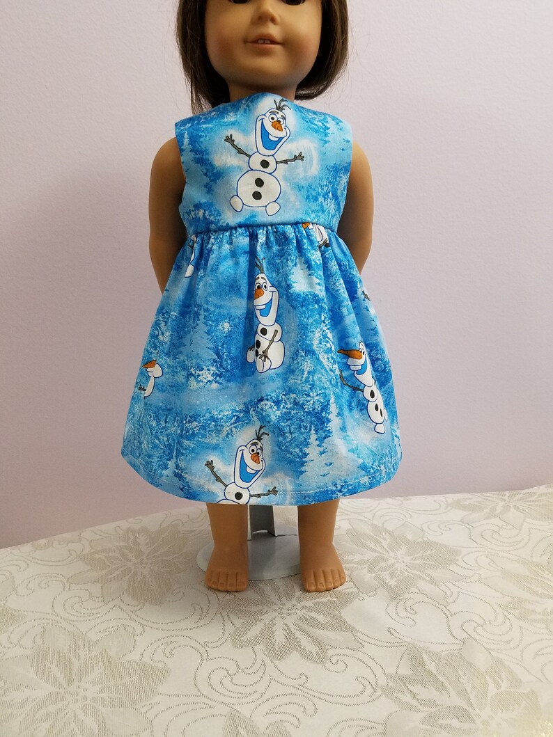 May include: A blue doll's dress with a pattern of Olaf the snowman from Frozen. The dress has a gathered skirt and a light blue and white winter forest background.